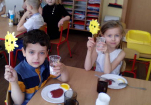 Dziewczynka i chłopiec podczas głosowania na smaczniejszy deser. Oboje unoszą deser owsiany i emblematy słoneczek na patyczkach w górę.